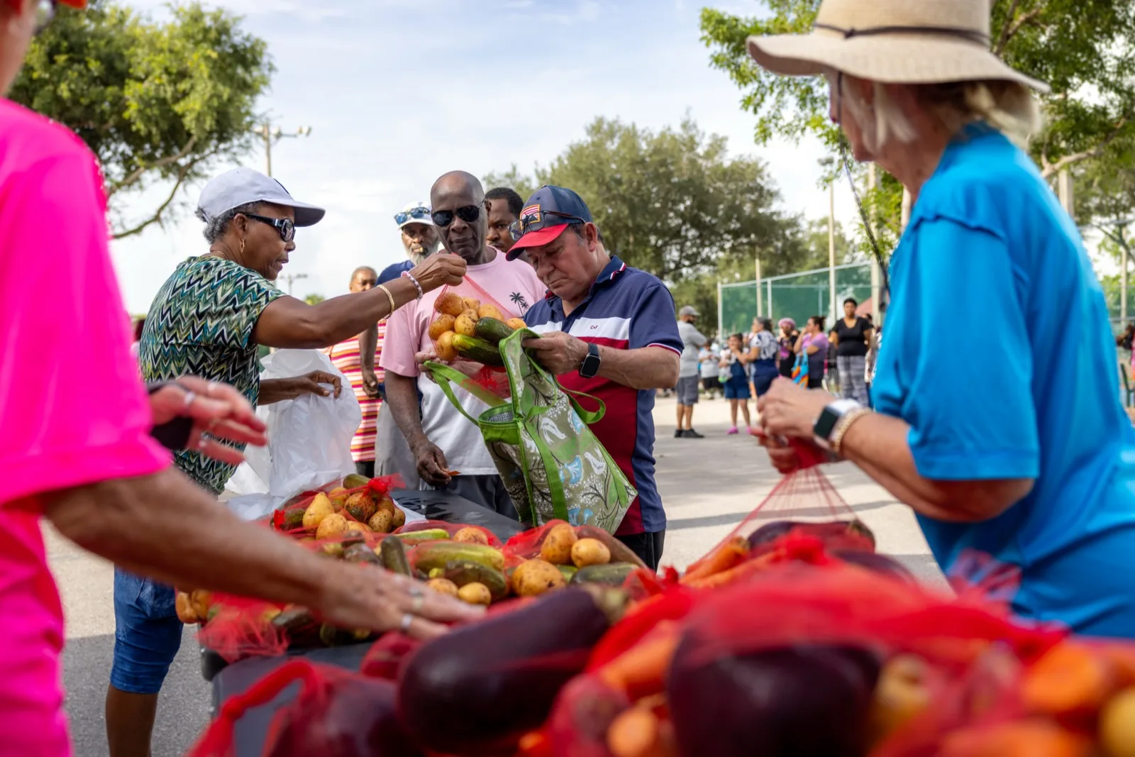 Volunteers and neighbors at an outdoor produce distribution, with bags of fresh vegetables on tables in the foreground