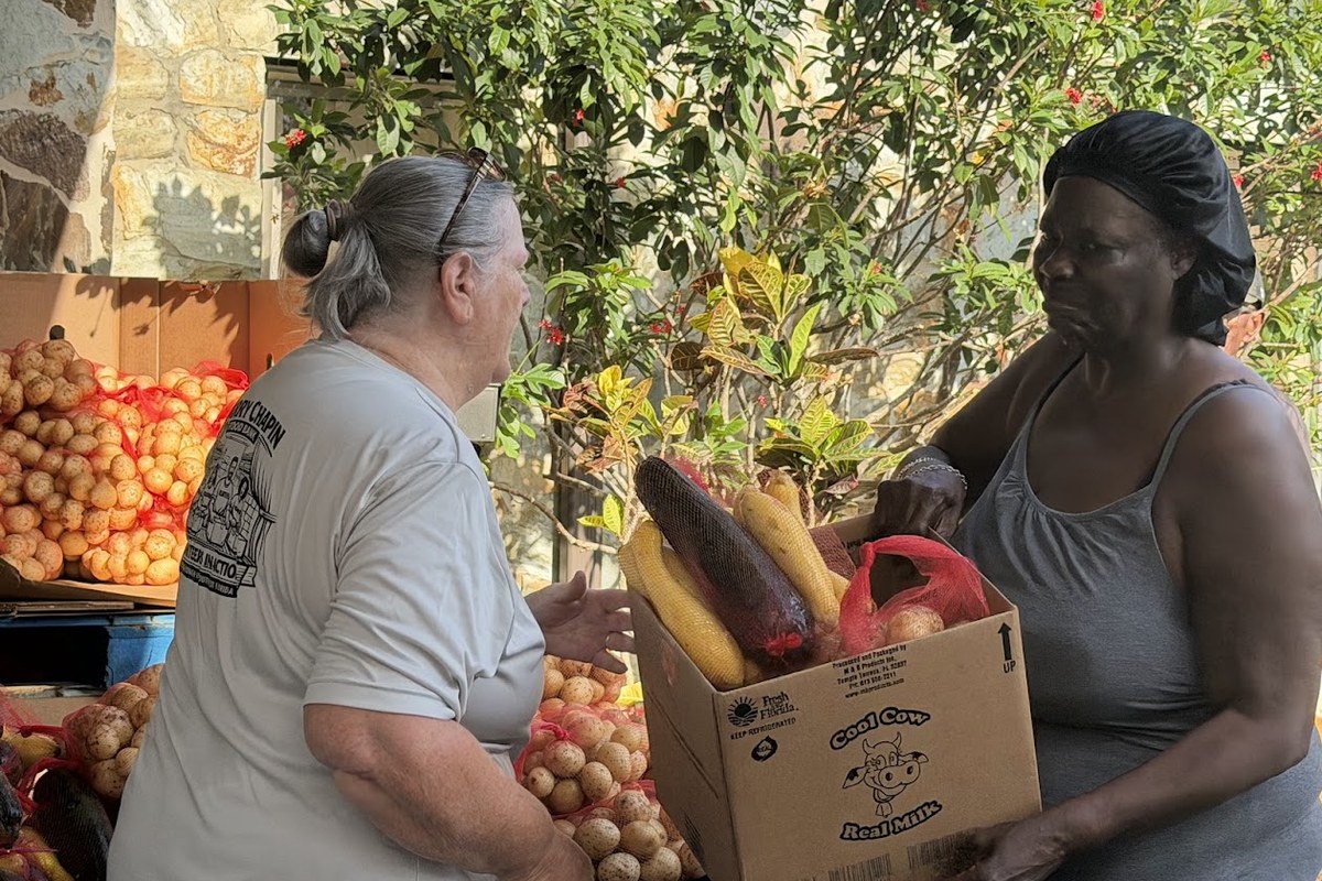 Debra, wearing a dark head covering and gray tank top, receives a box of corn and other fresh produce from a volunteer in a Harry Chapin Food Bank shirt, surrounded by crates of potatoes and citrus.