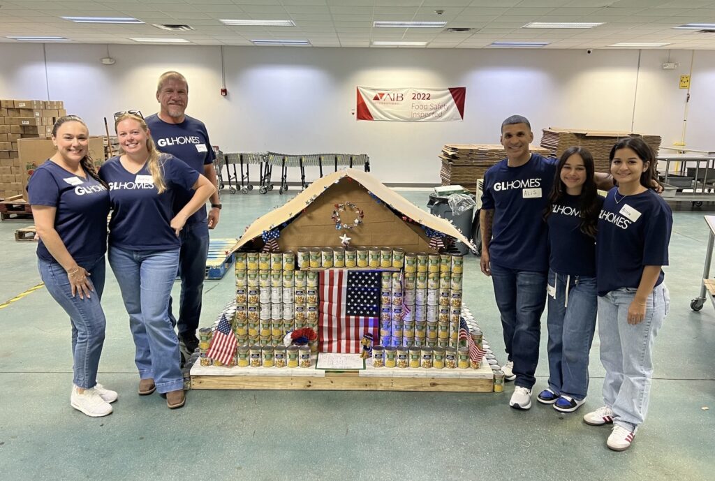 Six GLHomes volunteers stand beside a house-shaped display built from canned goods, decorated with American flags, at a Harry Chapin Food Bank food drive.