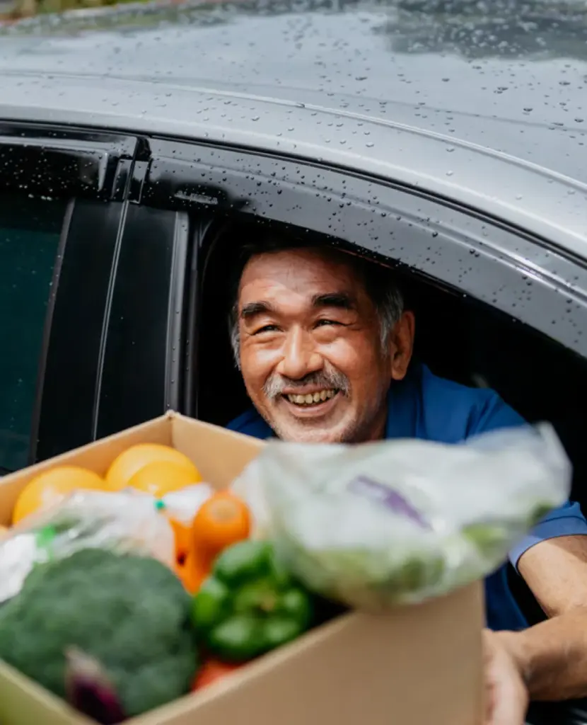 A smiling man receives a box of fresh produce through his car window