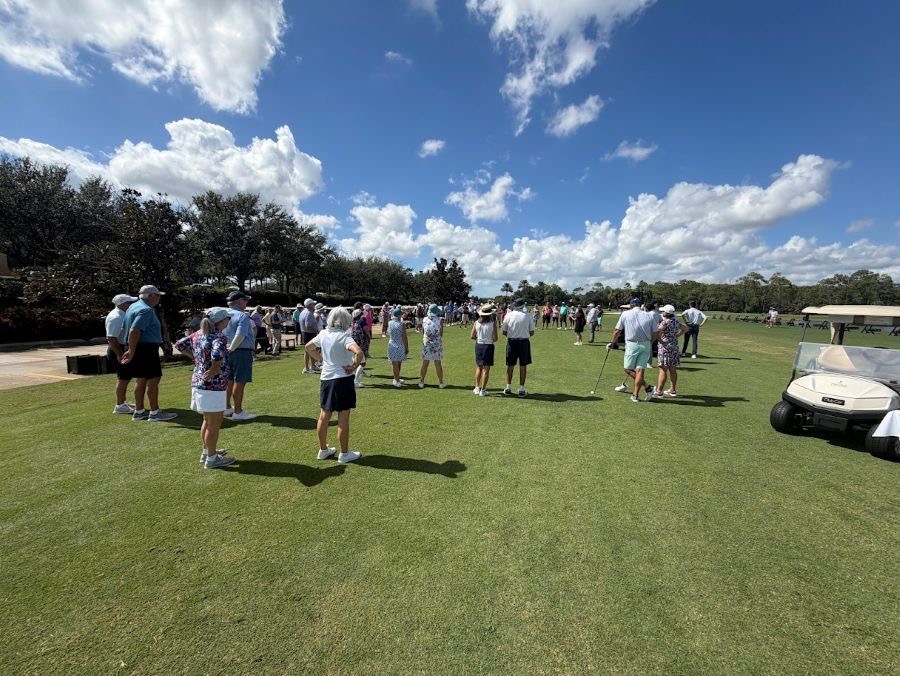 A group of golfers gathered on the fairway during the Heritage Cares Drive Out Hunger golf tournament, with golf carts and trees in the background under a blue sky.