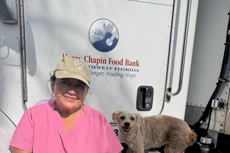 Maria, wearing a pink Harry Chapin Food Bank shirt and tan cap, smiles beside a small light-colored dog on the step of a Harry Chapin Food Bank truck.