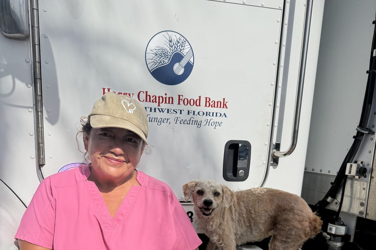 Maria, wearing a pink Harry Chapin Food Bank shirt and tan cap, smiles beside a small light-colored dog on the step of a Harry Chapin Food Bank truck.