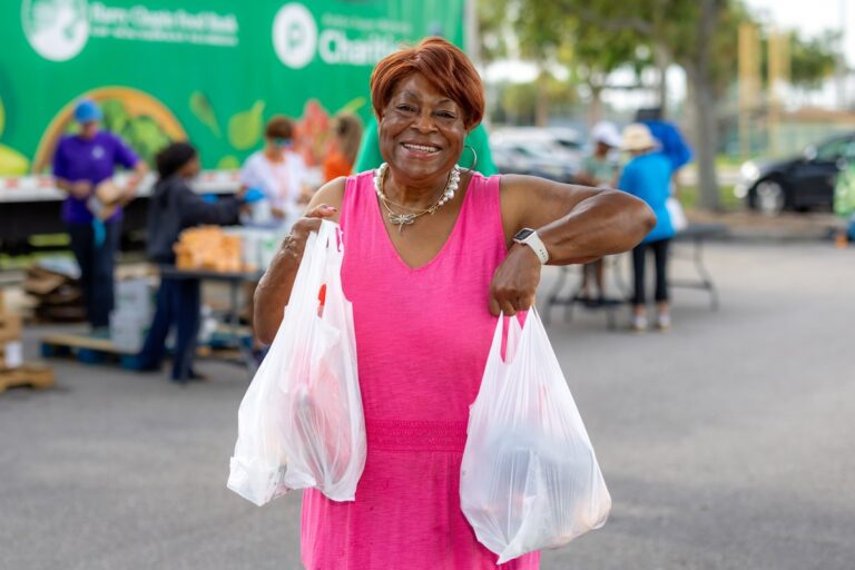 A smiling woman with red hair in a pink dress holds up two full plastic grocery bags in front of a Harry Chapin Food Bank branded truck and tent at an outdoor food distribution.