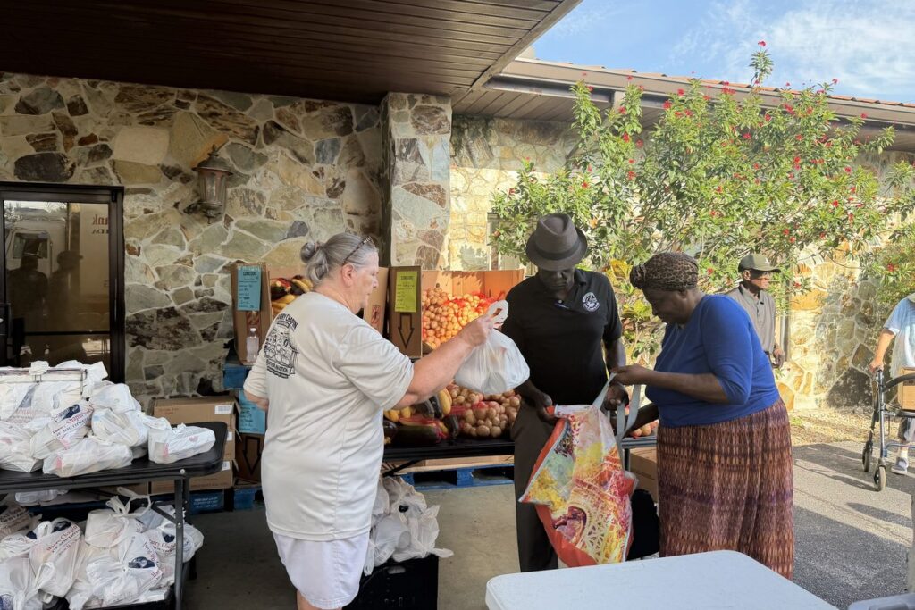 A volunteer hands a bag to two neighbors picking up fresh produce and groceries under a covered outdoor distribution area.