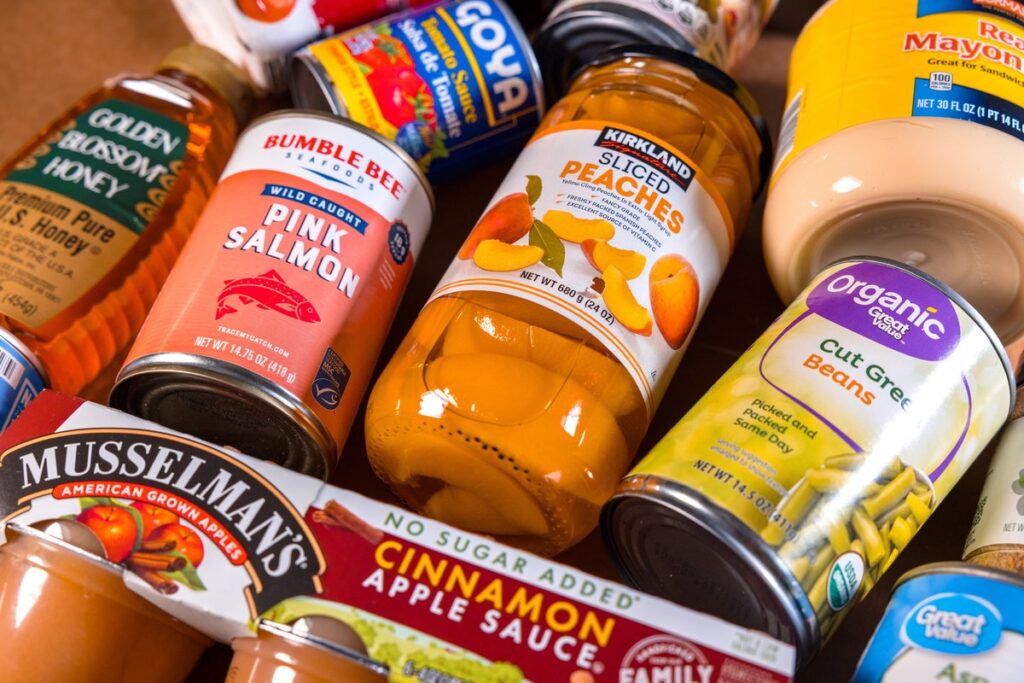 An assortment of canned and packaged pantry staples -- honey, canned salmon, peaches, green beans, applesauce -- packed closely together.
