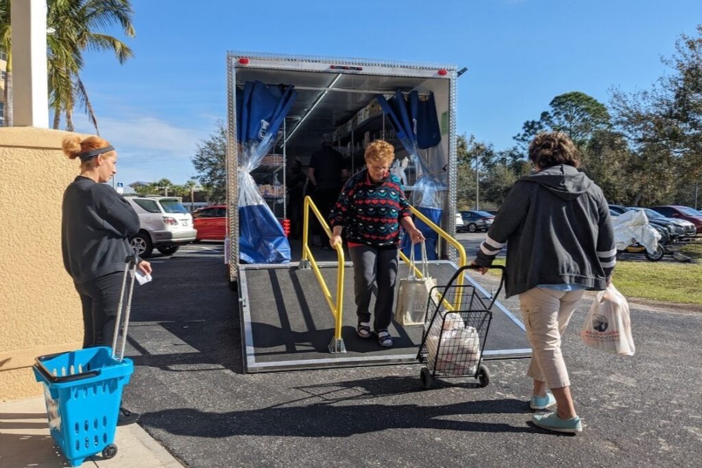 Three adults with shopping carts and grocery bags at the ramp of a mobile market trailer in a parking lot.