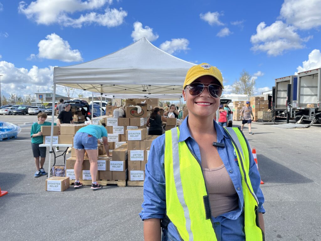 A smiling woman in a high-visibility vest at an outdoor food distribution.