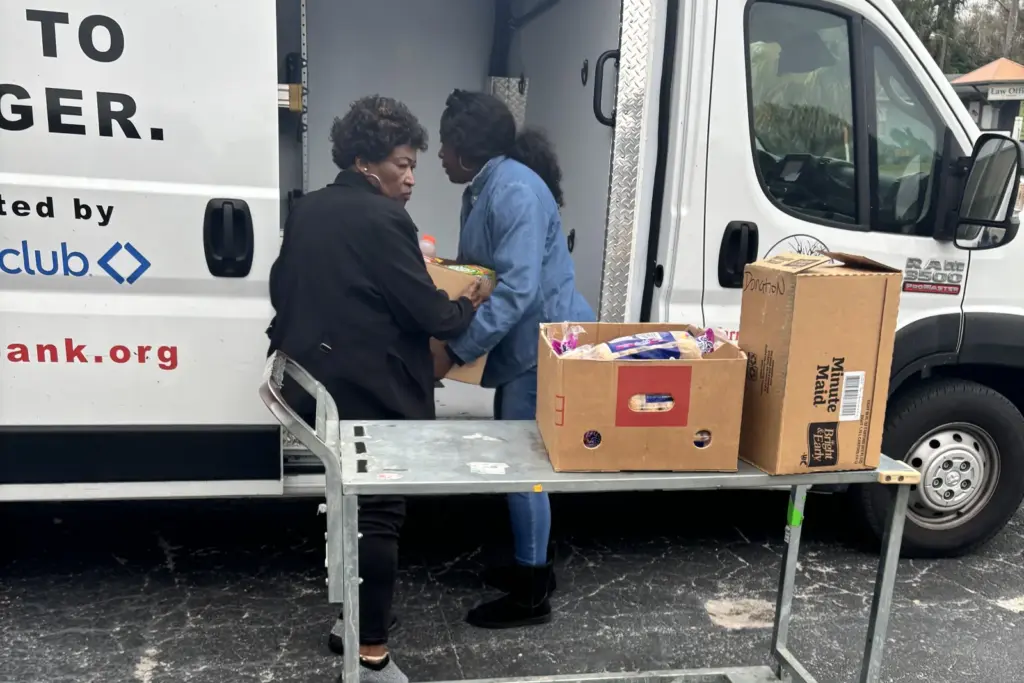 Two people from an agency partner lift boxes of donated groceries from a refrigerated van onto a metal cart at a retail store.