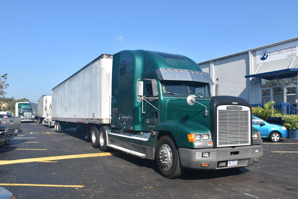 A green tractor-trailer parked outside a Harry Chapin Food Bank building, with more trucks lined up behind it.