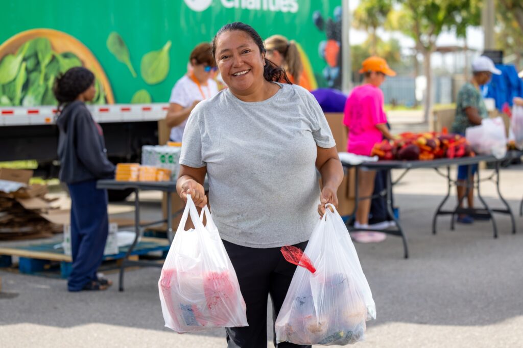 A smiling woman carries two full bags of groceries at an outdoor mobile food distribution, with volunteers at tables of fresh produce behind her.